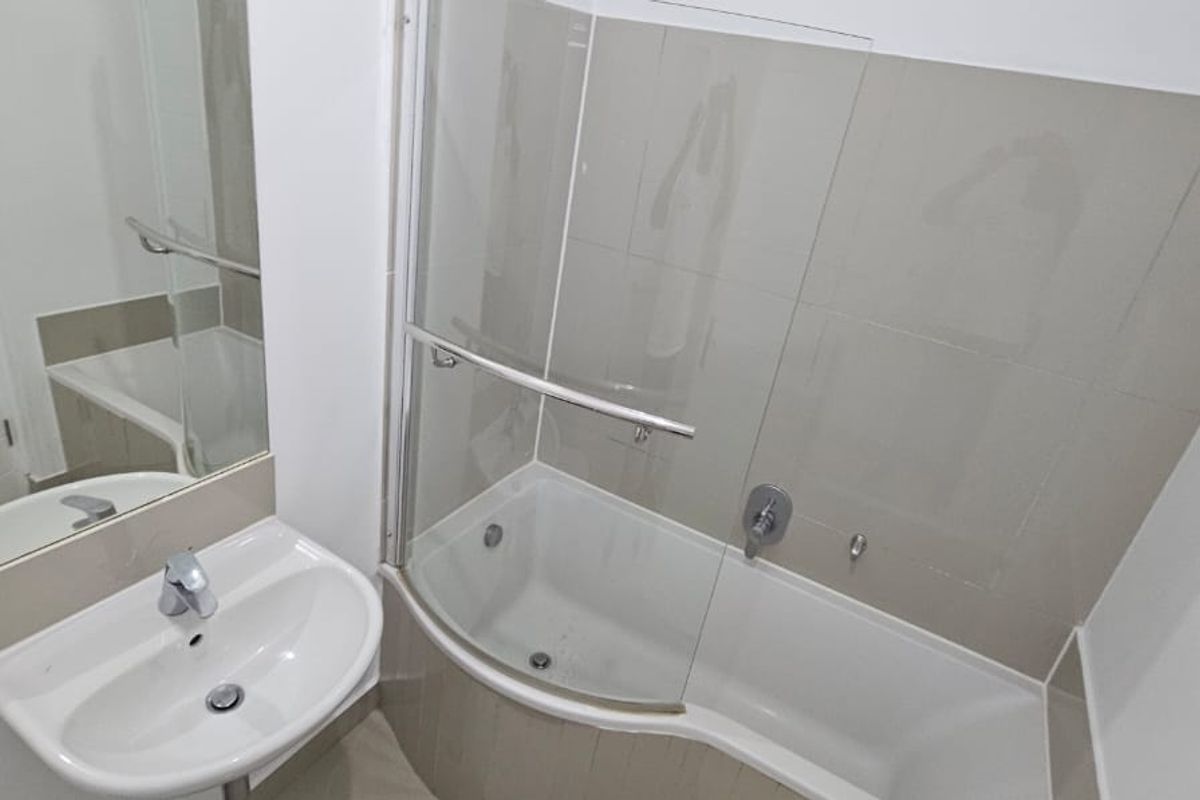 A clean, modern bathroom featuring a pedestal basin, wall-mounted mirror, and a shower-over-bath combination with a glass screen and neutral tiling.