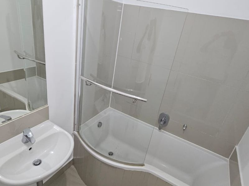 A clean, modern bathroom featuring a pedestal basin, wall-mounted mirror, and a shower-over-bath combination with a glass screen and neutral tiling.