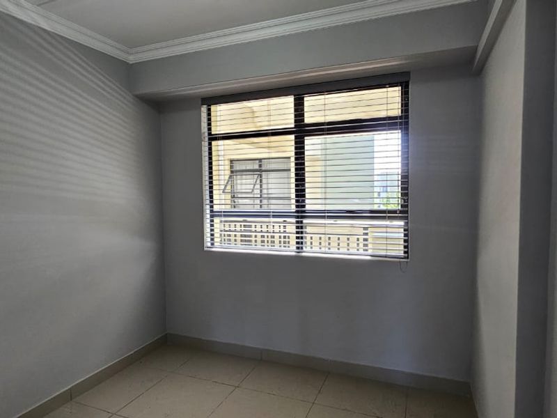 A clean, empty bedroom featuring neutral grey walls, tiled flooring, crown molding, and fitted venetian blinds.