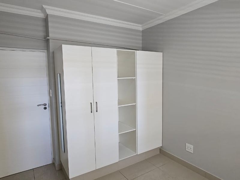 A clean, neutral bedroom featuring tiled flooring, crown molding, and a white built-in wardrobe with open shelving.