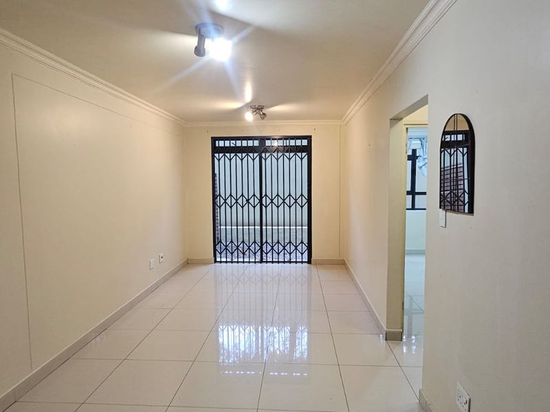 A bright, empty living area featuring neutral tiled flooring, ceiling cornices, and a secure metal gate leading to an outdoor space.