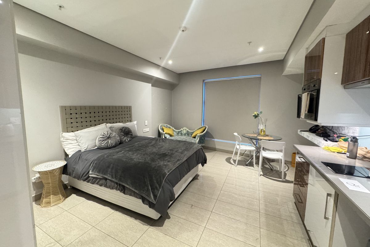 A modern open-plan studio apartment featuring a combined bedroom and kitchen area with sleek cabinetry, integrated appliances, and neutral tiled flooring.