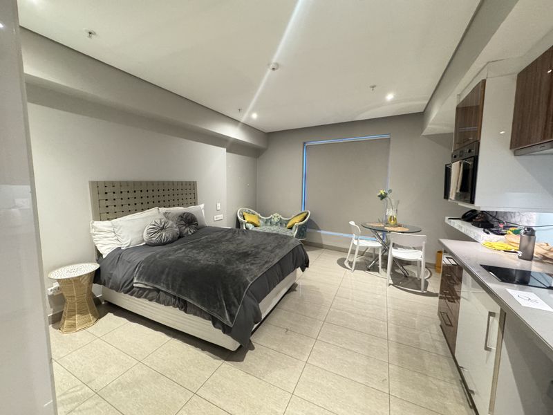 A modern open-plan studio apartment featuring a combined bedroom and kitchen area with sleek cabinetry, integrated appliances, and neutral tiled flooring.