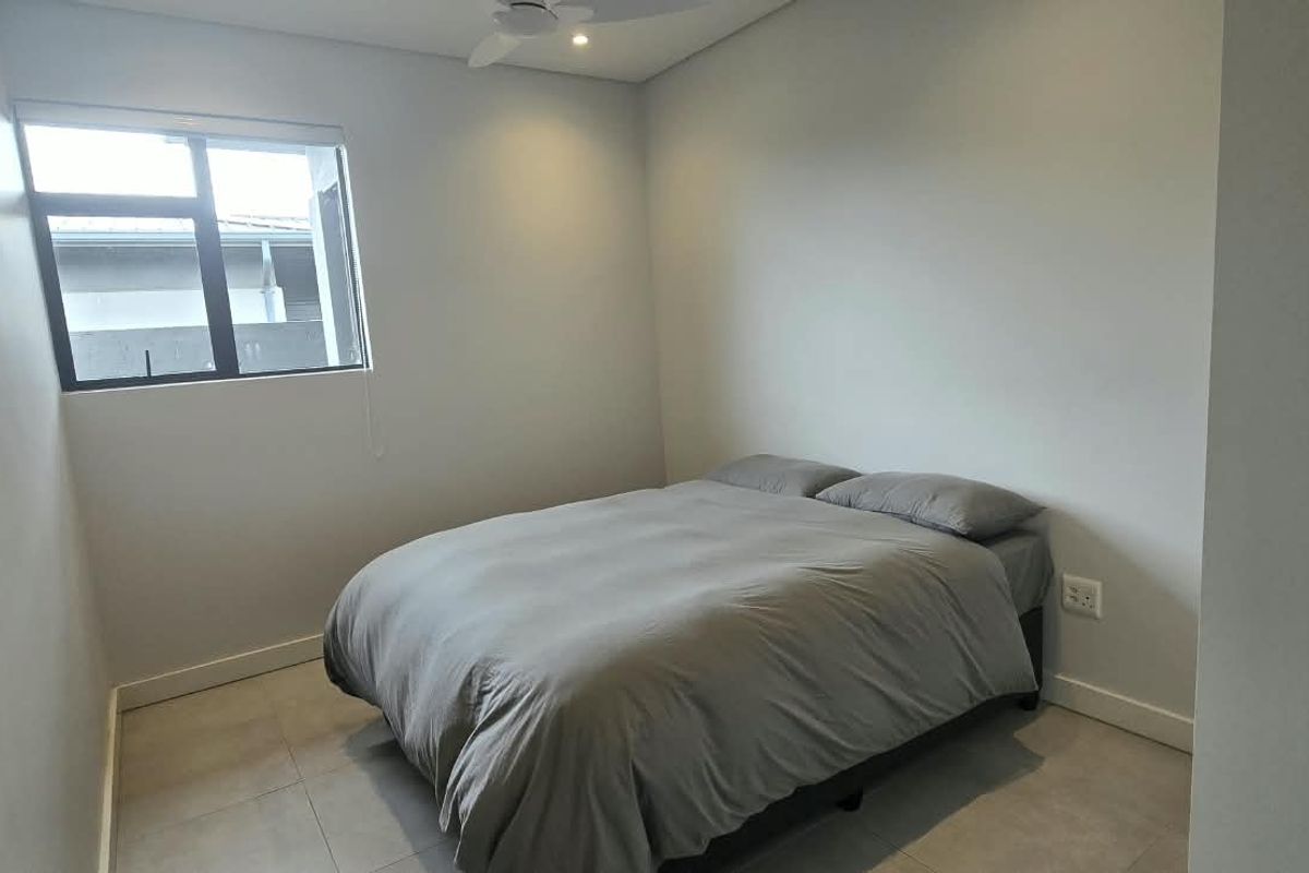 A simple bedroom featuring neutral tiled flooring, a ceiling fan, and an aluminium-framed window, providing a clean and functional living space.