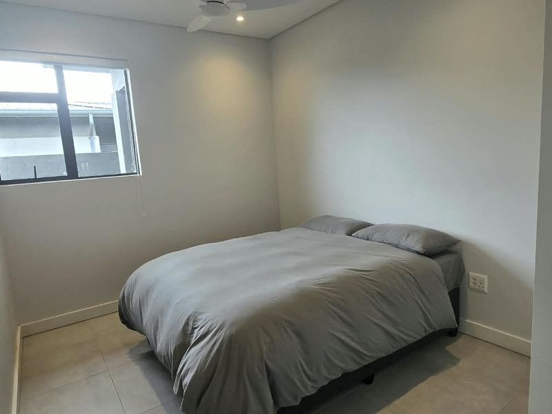 A simple bedroom featuring neutral tiled flooring, a ceiling fan, and an aluminium-framed window, providing a clean and functional living space.