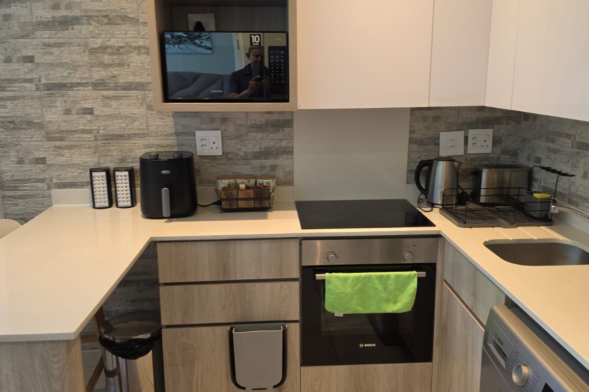 Modern compact kitchen featuring light wood-grain cabinetry, stone-look wall cladding, stone countertops, and integrated appliances including an oven and hob.