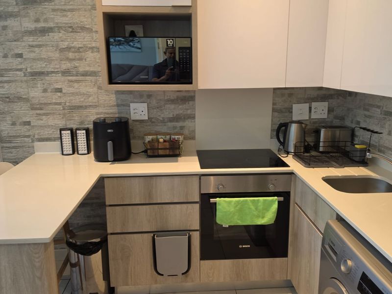 Modern compact kitchen featuring light wood-grain cabinetry, stone-look wall cladding, stone countertops, and integrated appliances including an oven and hob.