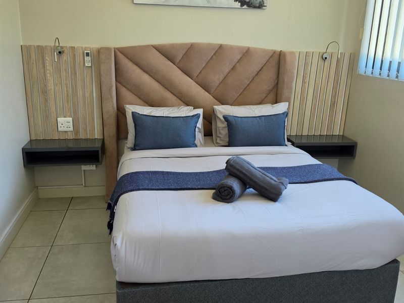A well-maintained bedroom featuring a custom built-in headboard with integrated floating bedside tables, vertical slat wall paneling, and neutral tiled flooring.