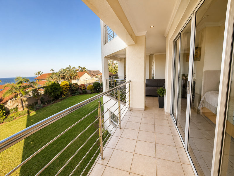 Spacious covered balcony featuring tiled flooring, stainless steel balustrades, and sliding glass door access, offering scenic views of the garden and ocean.