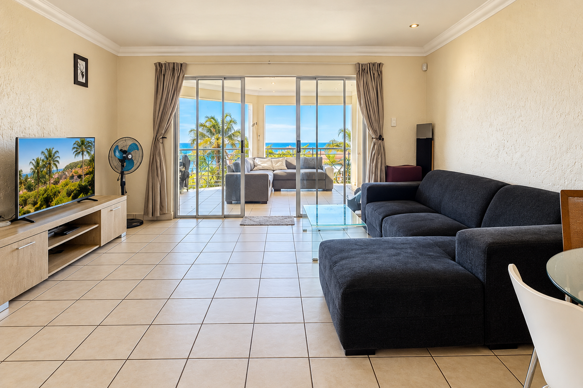 Spacious living room featuring neutral tiled flooring, coving, and large sliding glass doors that open onto a balcony with ocean views.