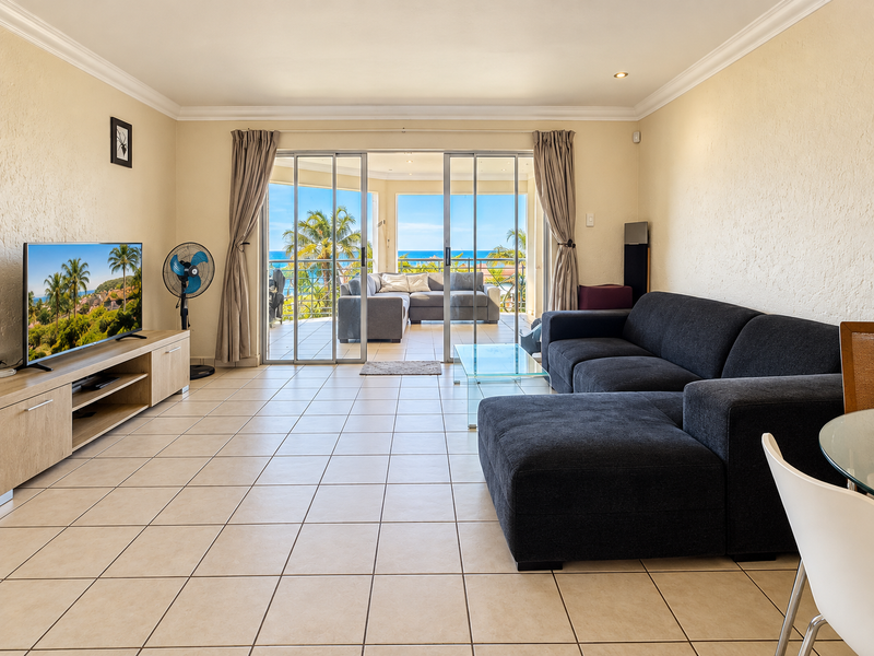 Spacious living room featuring neutral tiled flooring, coving, and large sliding glass doors that open onto a balcony with ocean views.