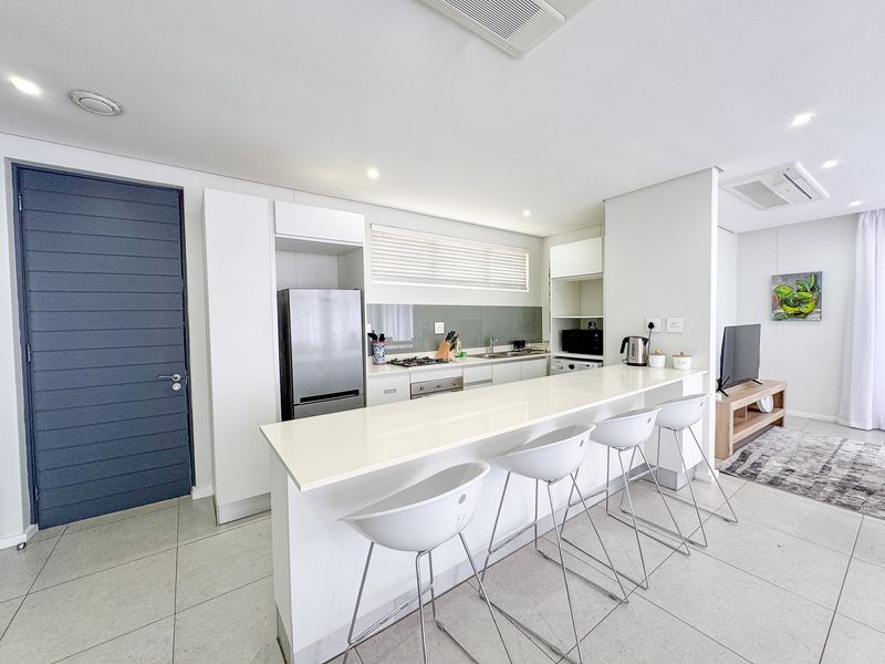 Modern open-plan kitchen featuring sleek white cabinetry, stone countertops, a breakfast bar, and integrated appliances, seamlessly flowing into the living area.