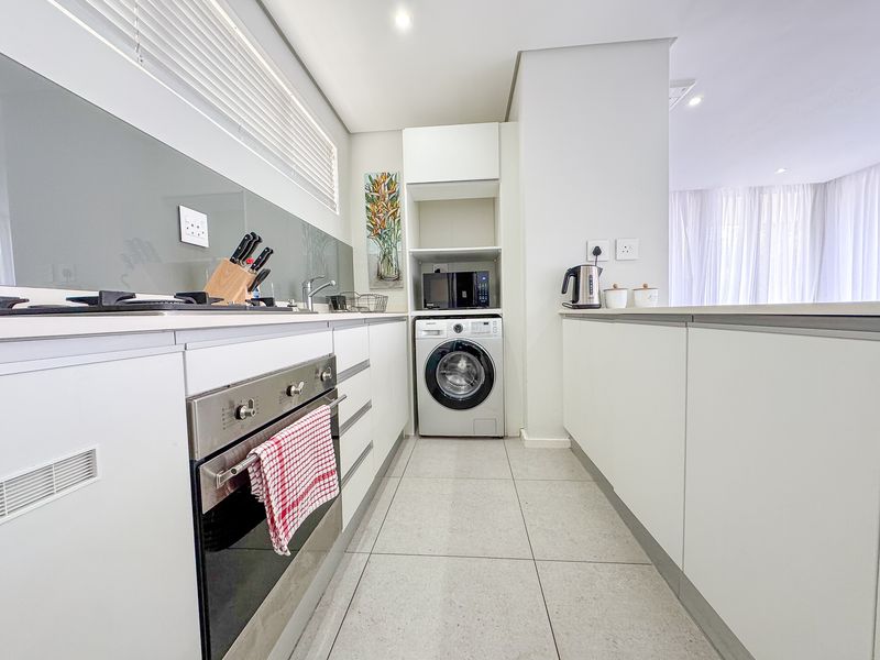 Modern galley-style kitchen featuring white cabinetry, stone countertops, integrated laundry space, built-in oven, and gas hob with a sleek glass splashback.