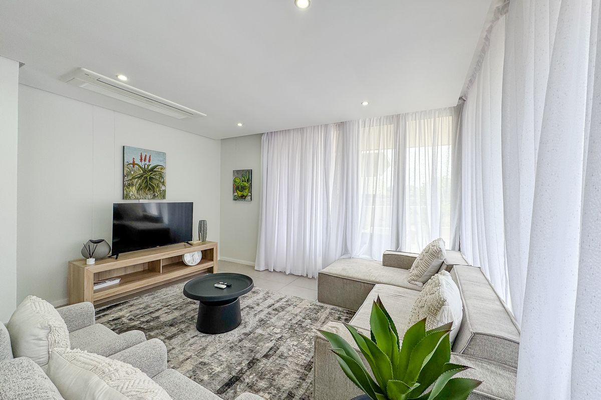 A bright and modern living room featuring tiled flooring, recessed downlighting, ducted air conditioning, and large windows with sheer curtains.