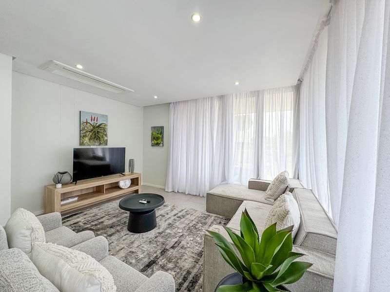 A bright and modern living room featuring tiled flooring, recessed downlighting, ducted air conditioning, and large windows with sheer curtains.