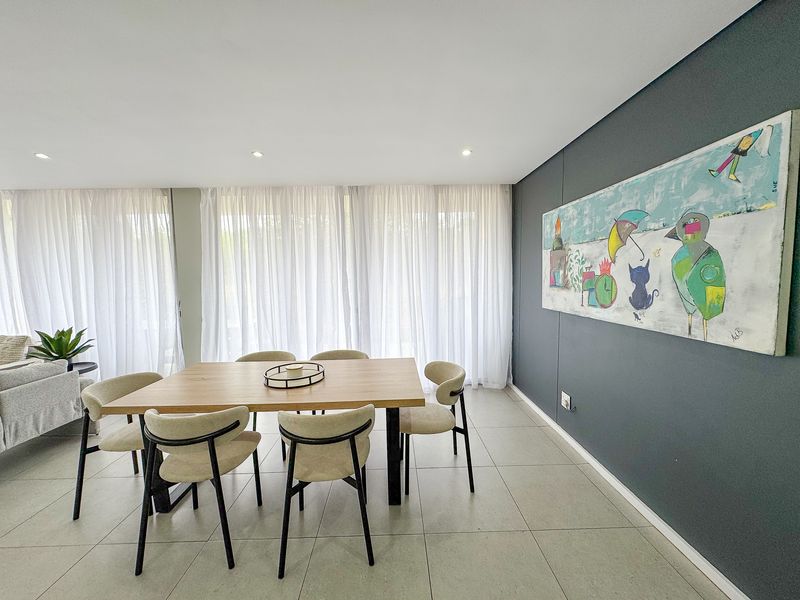 A bright and modern dining area featuring large windows with floor-to-ceiling sheer curtains, recessed lighting, and neutral tiled flooring.