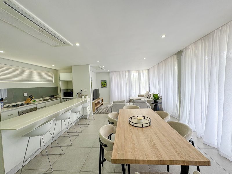 Modern open-plan living area featuring a sleek kitchen with stone island, integrated appliances, tiled floors, and floor-to-ceiling sheer curtains providing ample natural light.
