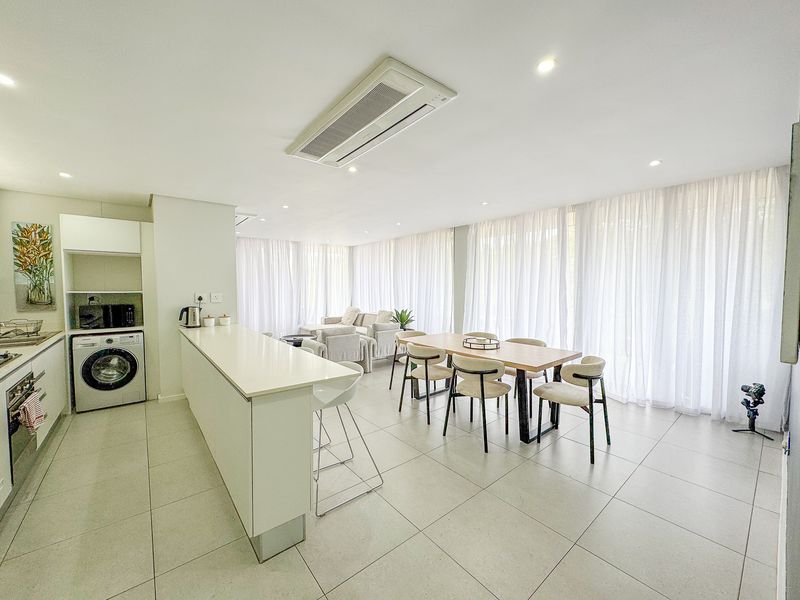 Modern open-plan living area featuring a sleek kitchen with integrated appliances, tiled flooring, ducted air conditioning, and floor-to-ceiling sheer curtains.