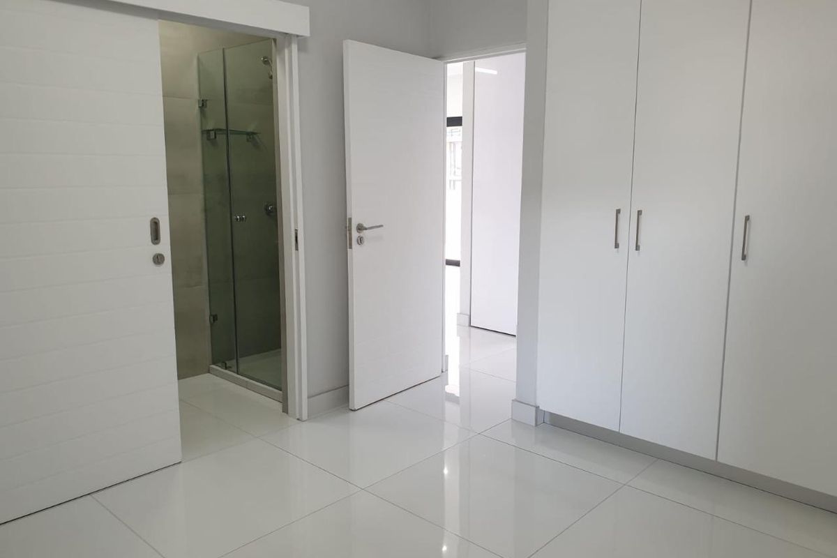 Modern bedroom featuring high-gloss built-in cupboards, polished tiled flooring, and direct access to a contemporary en-suite bathroom with a glass shower enclosure.