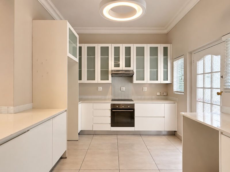 A clean, modern kitchen featuring white cabinetry, stone countertops, integrated oven and hob, and tiled flooring with ample natural light.