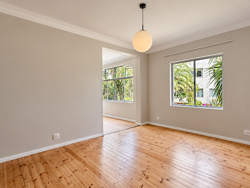 A bright, empty living room featuring polished timber flooring, crown molding, and a modern pendant light, with views of surrounding greenery.