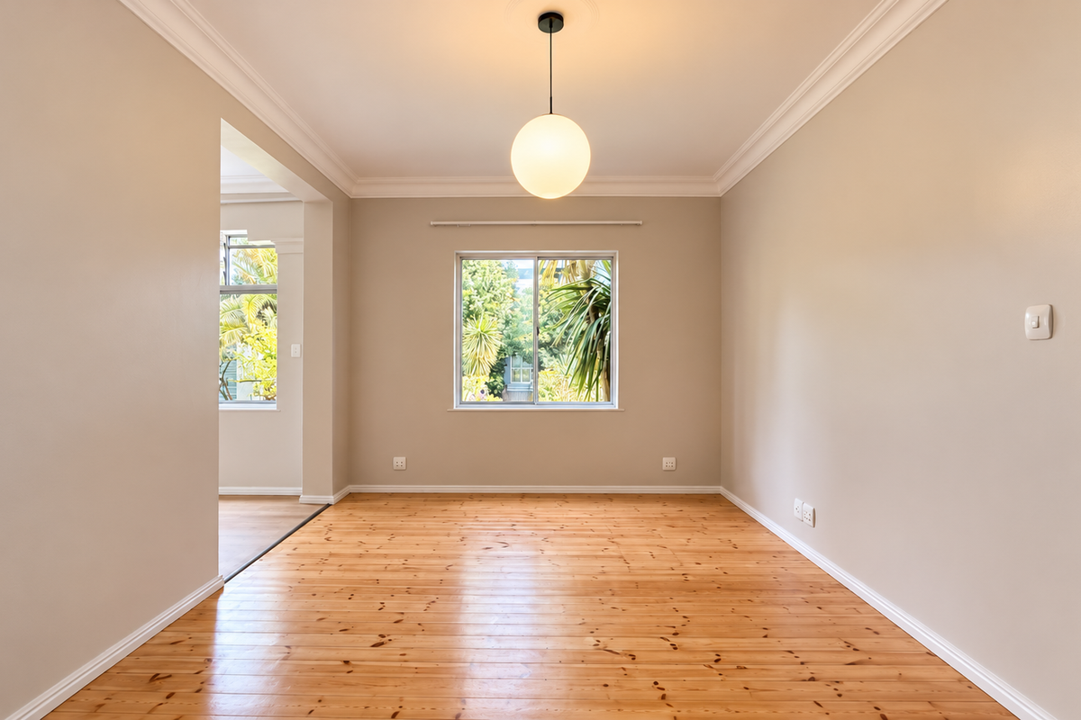 A bright, empty living room featuring polished timber flooring, crown molding, and a modern pendant light fixture with a view of the garden.
