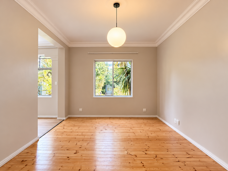 A bright, empty living room featuring polished timber flooring, crown molding, and a modern pendant light fixture with a view of the garden.