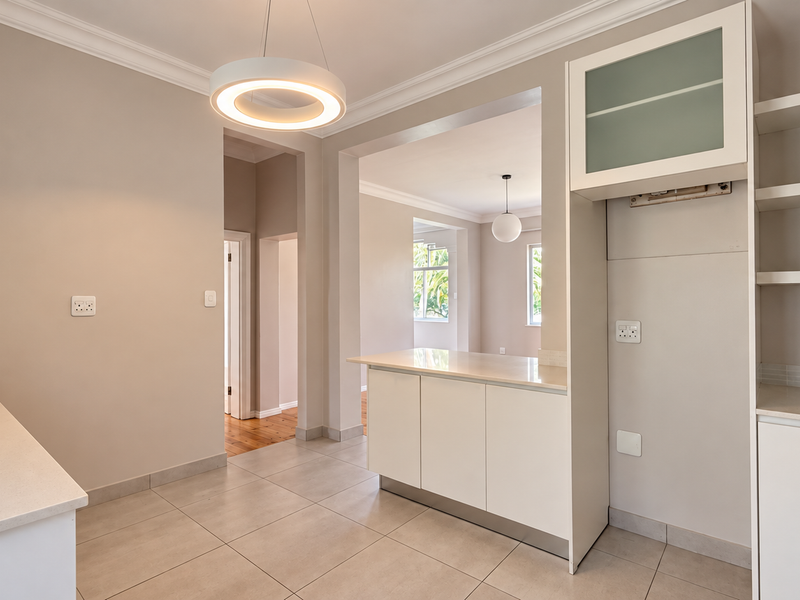 A bright, modern kitchen featuring clean white cabinetry, stone countertops, and tiled flooring, seamlessly flowing into the adjacent living area.