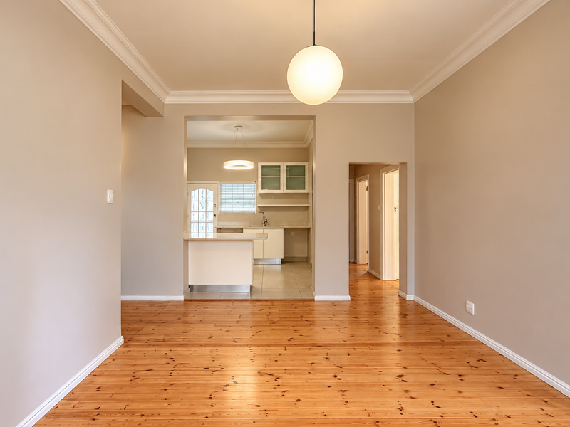 Spacious open-plan living area featuring polished timber flooring, crown molding, and a view through to a functional kitchen with built-in cabinetry.