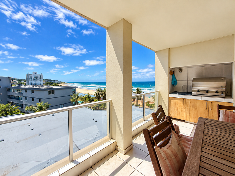 Covered balcony featuring a built-in braai, tiled flooring, and a glass balustrade, offering expansive ocean and coastal views.