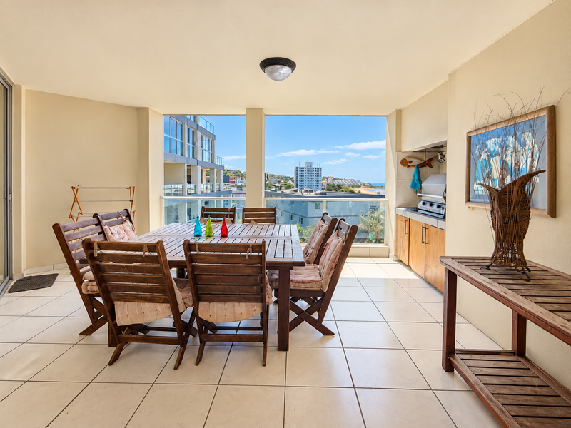Spacious covered balcony featuring a built-in braai, tiled flooring, and a glass balustrade with scenic urban and coastal views.