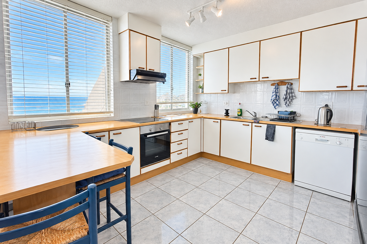A functional kitchen featuring white cabinetry, laminate countertops, tiled flooring, and integrated appliances, with large windows offering ocean views.