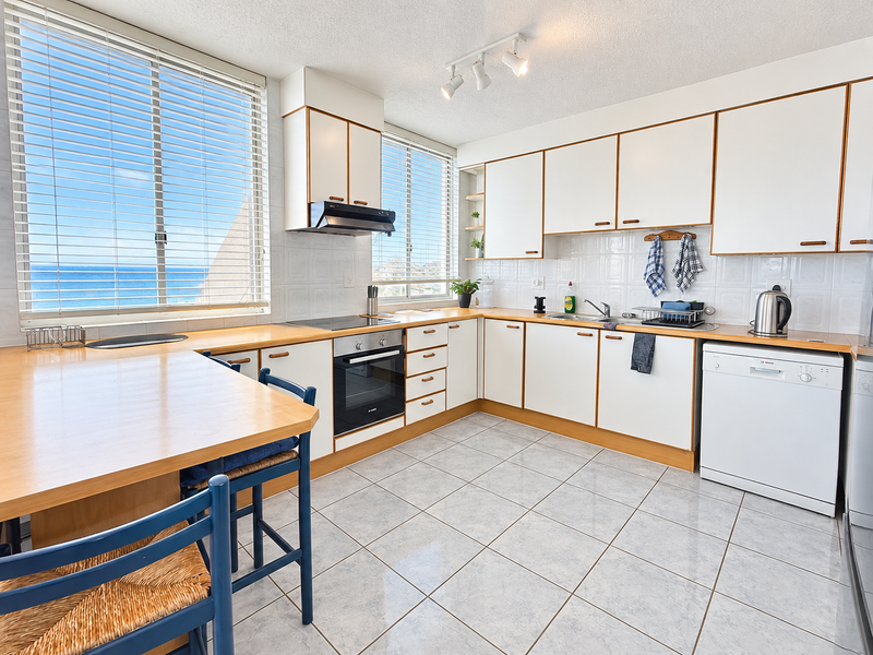 A functional kitchen featuring white cabinetry, laminate countertops, tiled flooring, and integrated appliances, with large windows offering ocean views.