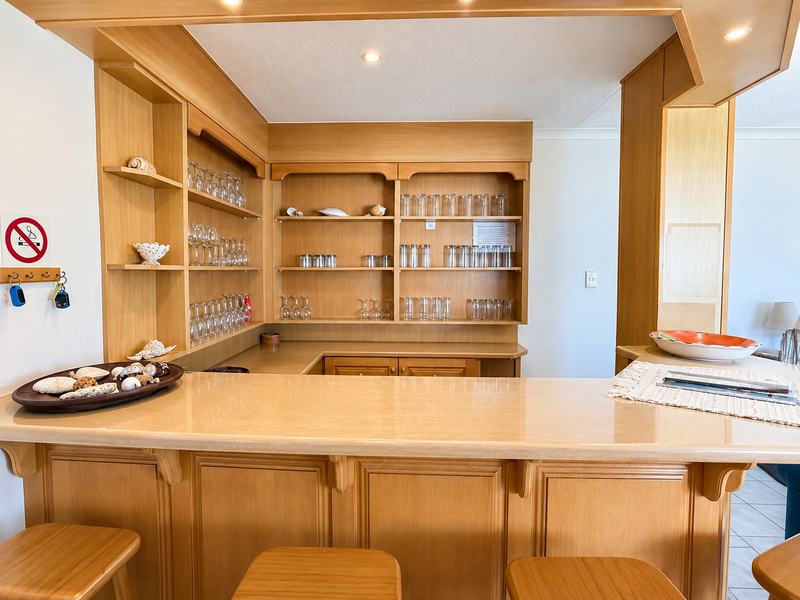A built-in wooden bar area featuring a stone countertop, integrated shelving, and recessed ceiling lighting, providing a functional space for entertaining.