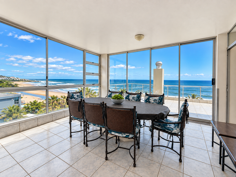 A bright, enclosed balcony featuring large glass windows with panoramic ocean views, tiled flooring, and direct access to an outdoor terrace area.