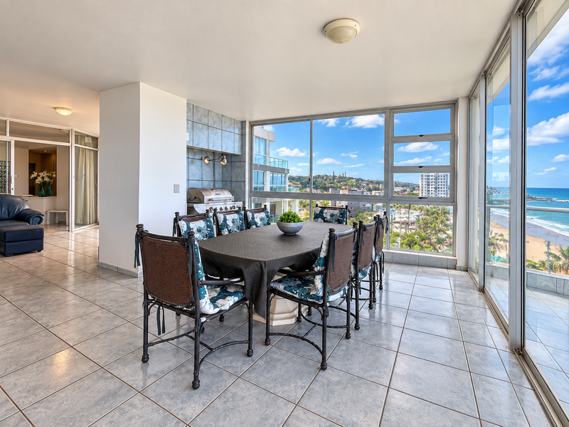 Enclosed balcony featuring tiled flooring, built-in braai, and expansive glass windows offering panoramic ocean and coastal views.