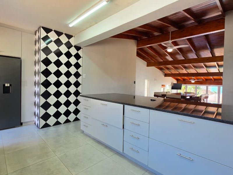 Spacious open-plan kitchen featuring modern cabinetry, granite countertops, and a distinctive checkered feature wall, flowing into a living area with exposed timber beams.