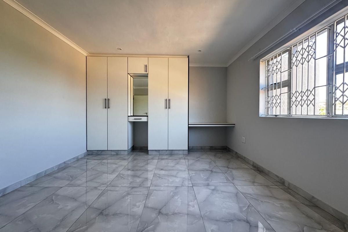 A bright, empty bedroom featuring modern grey tiled flooring, built-in cupboards with an integrated vanity, and a built-in desk under a window with security bars.