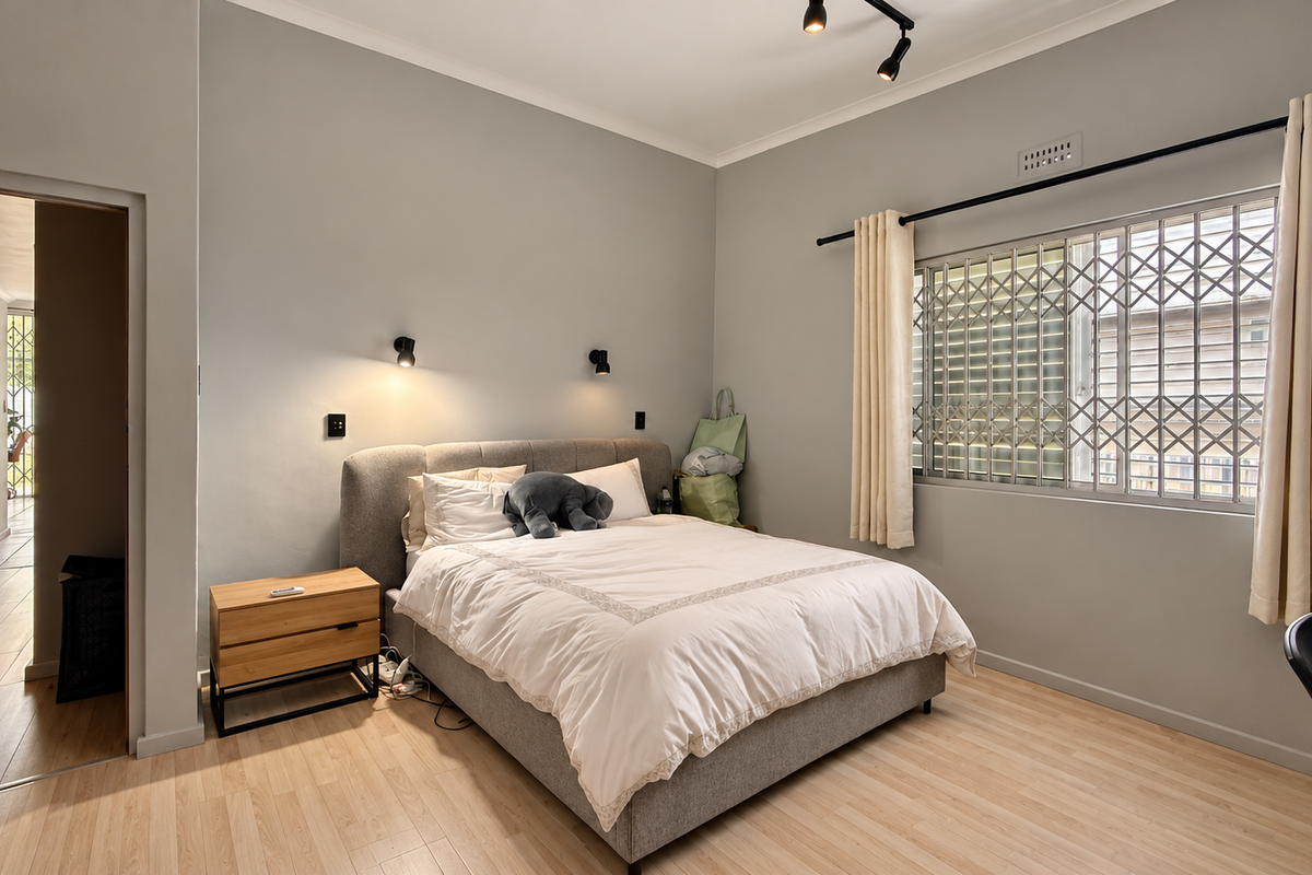 A well-lit bedroom featuring light laminate flooring, neutral grey walls, and a secure window with an integrated security gate.