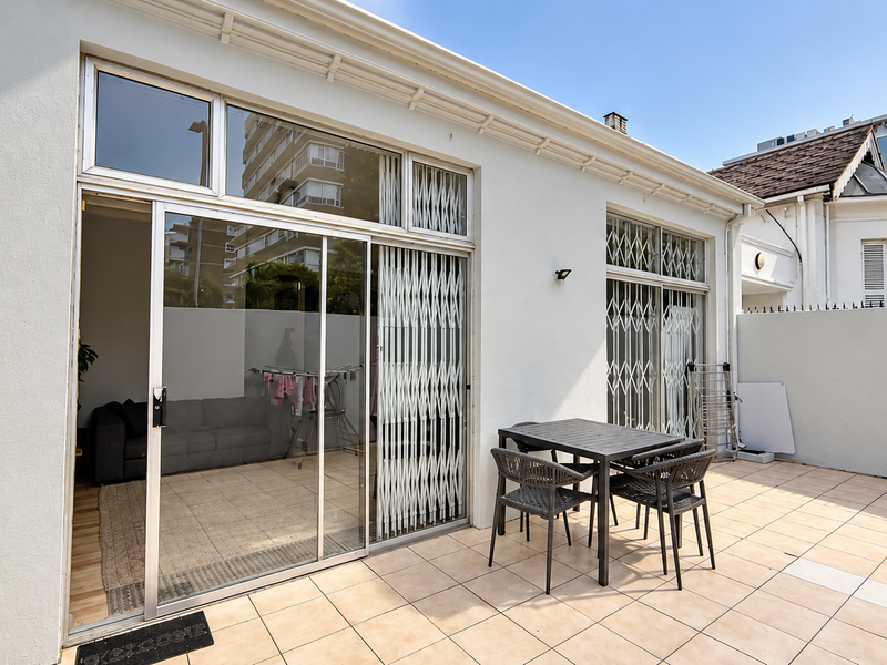 A paved outdoor patio area featuring sliding glass doors with security gates, providing seamless access to the interior living space.