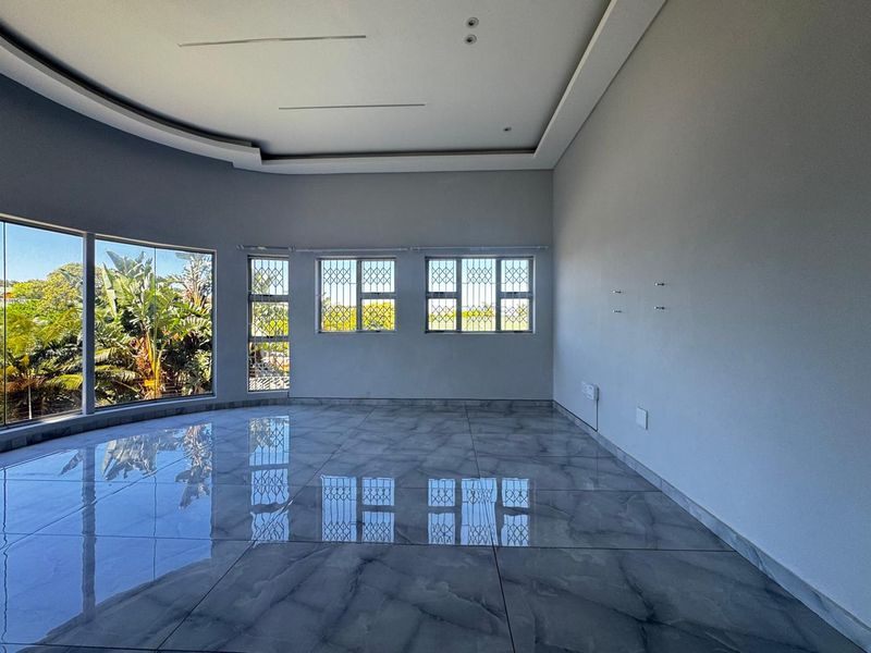 A bright, empty living room featuring polished porcelain floor tiles, recessed ceiling detail, and large windows with security bars overlooking the garden.