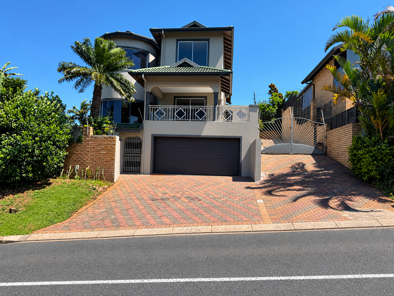 Front elevation of a multi-story residential house featuring a paved driveway, double garage, and an upper-level balcony with metal railings.
