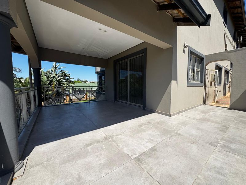 A spacious covered balcony featuring modern tiled flooring, a decorative metal balustrade, and a sliding glass door entrance.