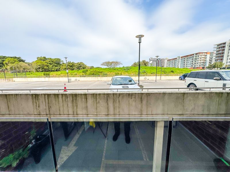 A balcony featuring a glass balustrade and concrete parapet wall, overlooking an open parking area and surrounding landscape.