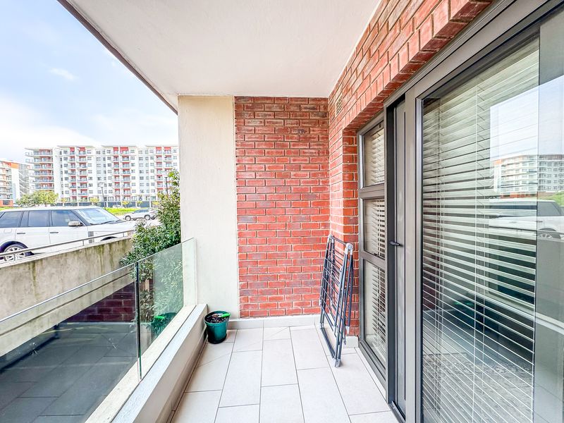A clean, covered balcony featuring modern tiled flooring, an exposed brick feature wall, and a glass balustrade overlooking the surrounding apartment complex.