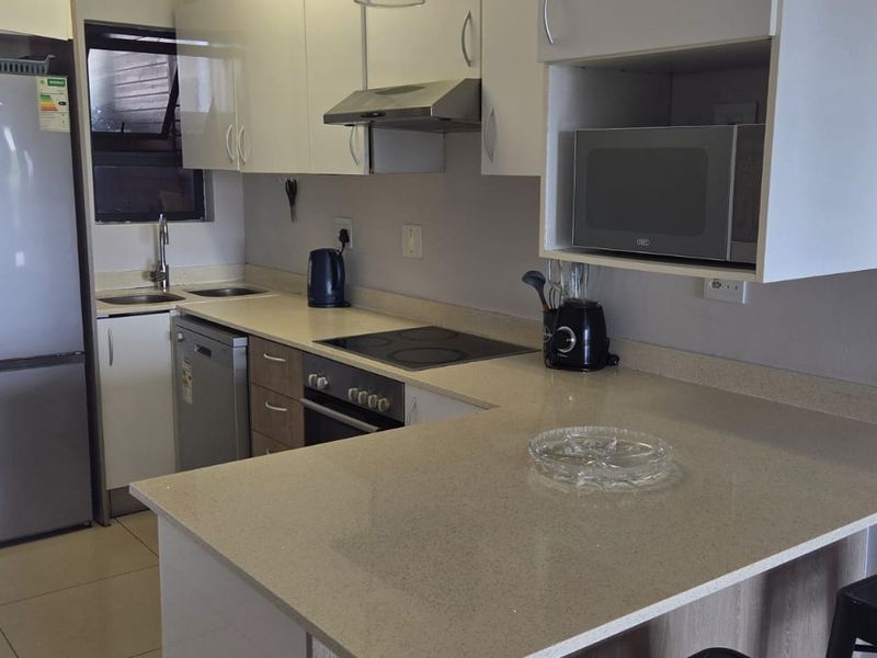 Modern kitchen featuring sleek white cabinetry, stone countertops, integrated appliances, and a breakfast bar, seamlessly connected to an outdoor balcony area.