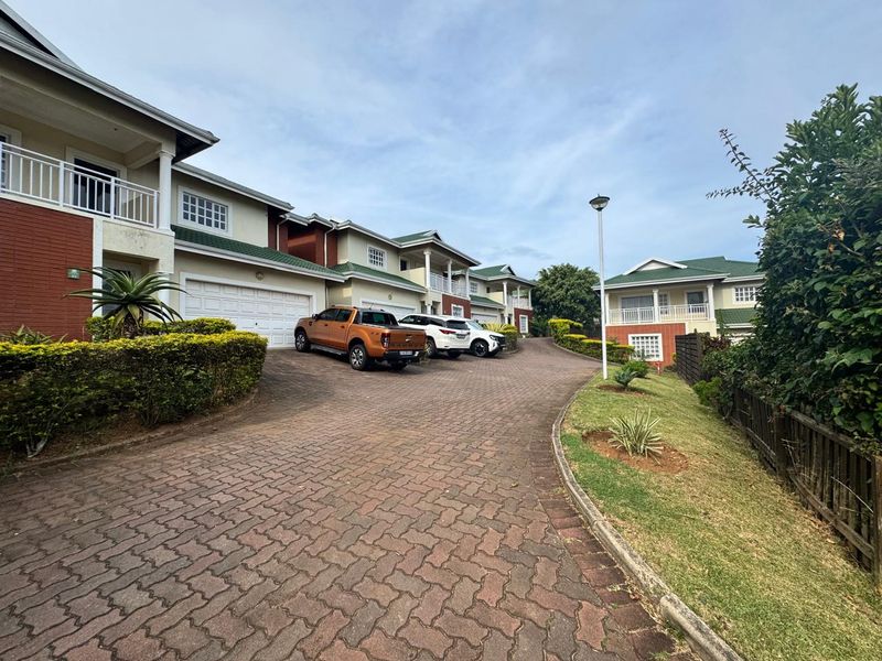 A view of the townhouse complex exterior featuring a paved driveway, multiple garage entrances, and upper-level balconies with a well-maintained garden area.