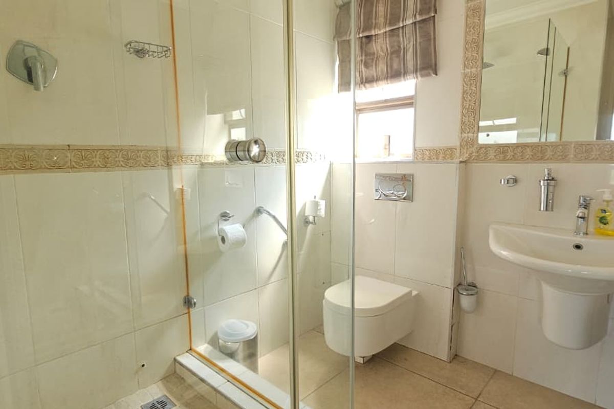 A functional bathroom featuring a glass-enclosed shower, wall-mounted toilet, and pedestal washbasin with neutral wall and floor tiling.