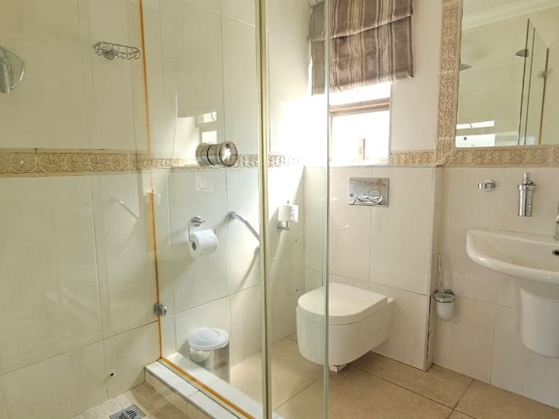 A functional bathroom featuring a glass-enclosed shower, wall-mounted toilet, and pedestal washbasin with neutral wall and floor tiling.