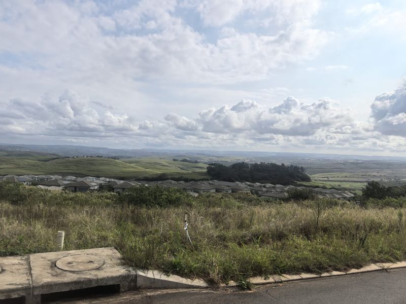 Elevated view overlooking a residential estate and surrounding rolling hills, featuring a foreground stormwater drain and natural vegetation.
